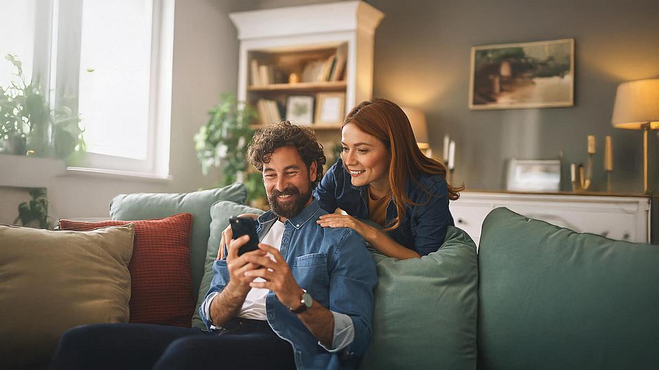 Glückliches Paar genießt die gemeinsame Zeit in ihrem neuen Zuhause, während sie auf einem gemütlichen Sofa ihre Smartphones erkunden