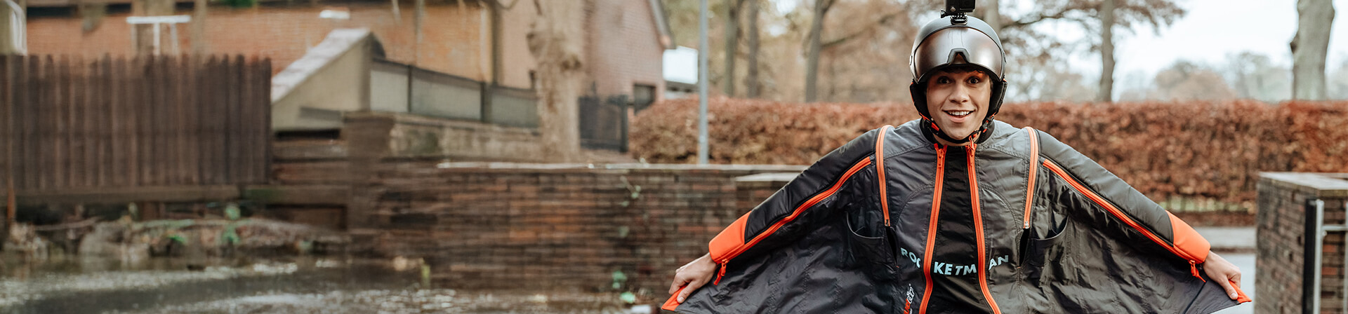 Lächelnde Person im Freestyle-Sprunganzug mit Helm und montierter Actionkamera, steht im Freien vor herbstlichen Bäumen und blickt direkt in die Kamera. Lächelnde Person im Freestyle-Sprunganzug mit Helm und montierter Actionkamera, steht im Freien vor herbstlichen Bäumen und blickt direkt in die Kamera.