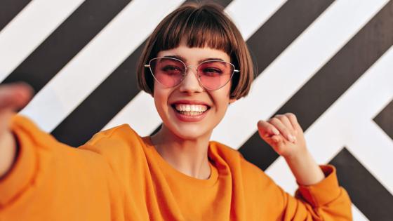 Frau mit Sonnenbrille lächelt in die Kamera vor einem gestreiften Hintergrund.